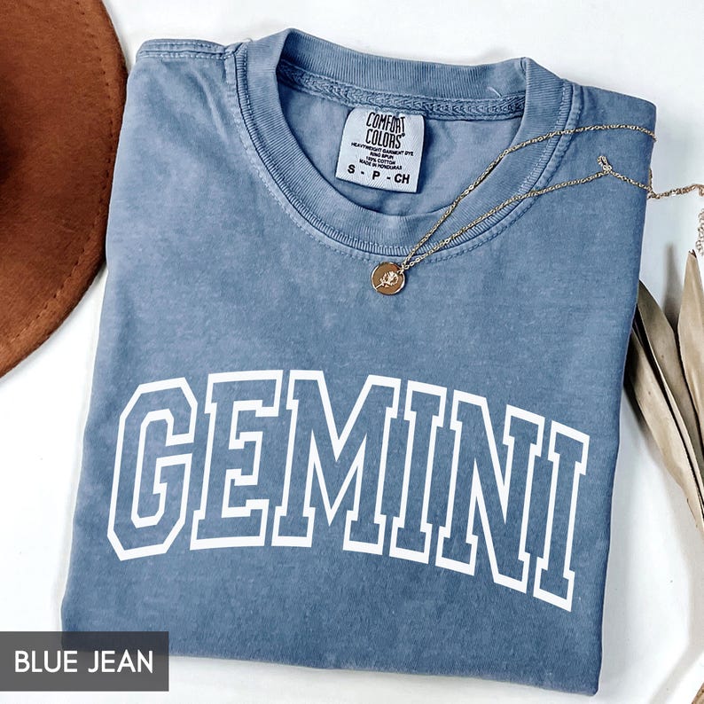 May include: Blue jean colored t-shirt with the word "GEMINI" in white block letters. The shirt has a crew neck and a small tag at the neckline. A gold necklace with a pendant is draped across the shirt.