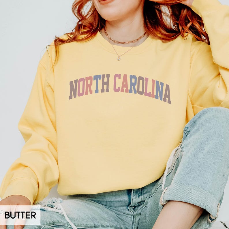 Puede incluir: Una sudadera amarilla clara con el texto "NORTH CAROLINA" en una fuente multicolor de estilo universitario. La parte superior de manga larga est&aacute; hecha de un material suave y es adecuada para ropa informal.