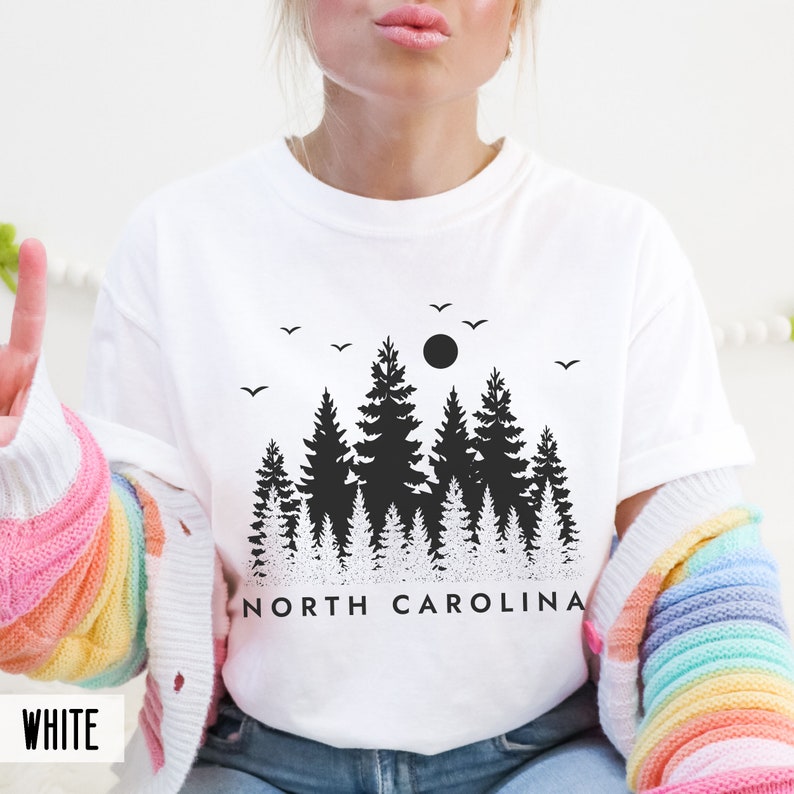 以下が含まれることがあります： 黒い森のシルエットと「NORTH CAROLINA」の文字が書かれた白のTシャツです。