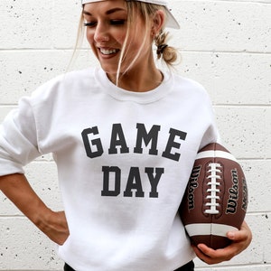 May include: A white sweatshirt with the text "GAME DAY" printed in black. A person is holding a brown and white football.