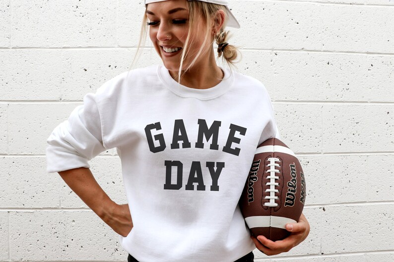 May include: A white sweatshirt with the text "GAME DAY" printed in black. A person is holding a brown and white football.