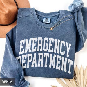May include: A denim blue sweatshirt with the words "EMERGENCY DEPARTMENT" in large white block letters. A gold necklace with a pendant is draped across the front. A brown hat is visible in the background.