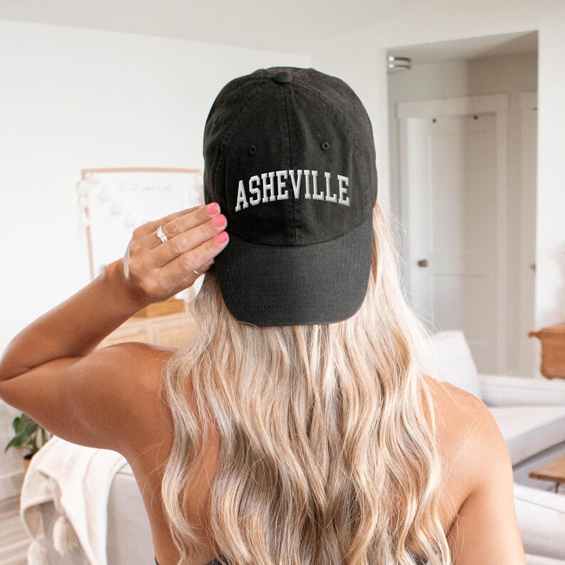 Puede incluir: Gorra de b&eacute;isbol negra con la palabra "ASHEVILLE" bordada en blanco. La gorra la lleva una persona con pelo largo y rubio. El fondo incluye un vistazo a una sala de estar.