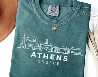 May include: A blue spruce t-shirt featuring a white line art illustration of the Athens, Greece skyline. The text "ATHENS GREECE" is printed in white below the skyline. The shirt has a Comfort Colors label on the inside collar.