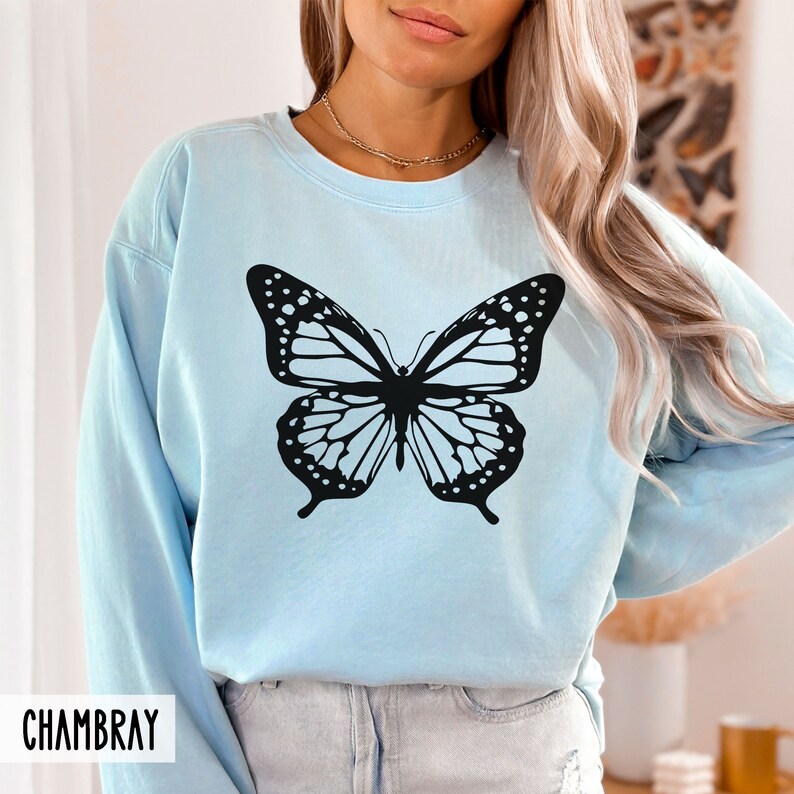 Peut inclure: Un sweat-shirt bleu clair avec un motif de papillon noir. Le sweat-shirt a un col rond et des manches longues. Le mot "CHAMBRAY" est imprim&eacute; dans le coin inf&eacute;rieur gauche de l'image.