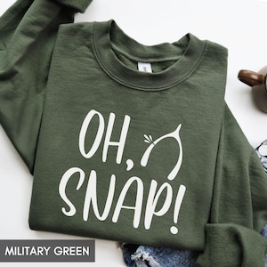 May include: Military green sweatshirt with the text "OH, SNAP!" in white script. The design includes a wishbone graphic. The sweatshirt is a crew neck style.