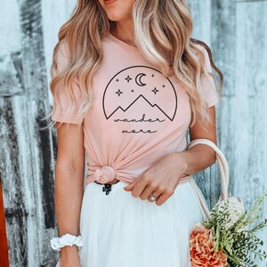 A woman with blonde-streaked hair wears sunglasses, a pink t-shirt with a black graphic, a white tulle skirt, and holds a bag of flowers.