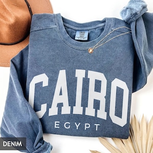 May include: A denim blue sweatshirt with the word "CAIRO" in large white block letters, with "EGYPT" below. A gold necklace with a pendant is draped on the sweatshirt. A brown hat is visible in the background.