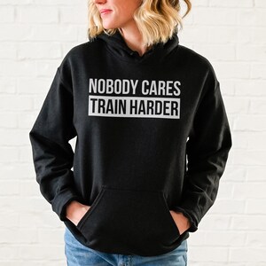 Pu&ograve; includere: Felpa con cappuccio nera con il testo "Nobody Cares Train Harder" in lettere bianche.