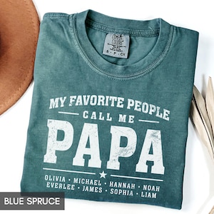 A green t-shirt with the text "MY FAVORITE PEOPLE CALL ME PAPA" printed on it, along with the names "OLIVIA, MICHAEL, HANNAH, NOAH, EVERLEE, JAMES, SOPHIA, LAM" printed below. The t-shirt is laid out on a white surface, with a brown hat and some leaves visible in the background.