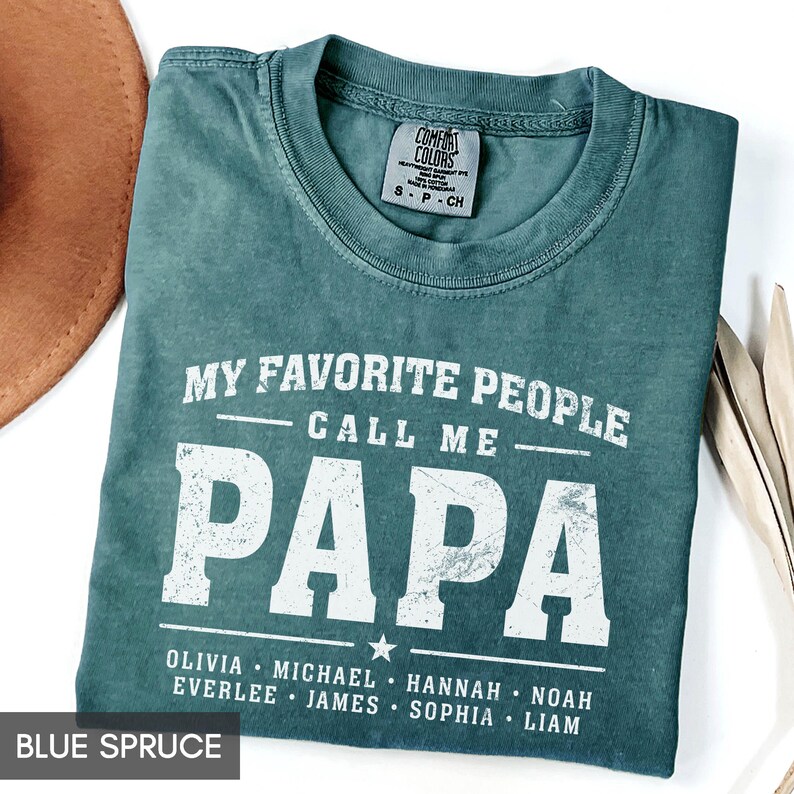 A green t-shirt with the text "MY FAVORITE PEOPLE CALL ME PAPA" printed on it, along with the names "OLIVIA, MICHAEL, HANNAH, NOAH, EVERLEE, JAMES, SOPHIA, LAM" printed below. The t-shirt is laid out on a white surface, with a brown hat and some leaves visible in the background.