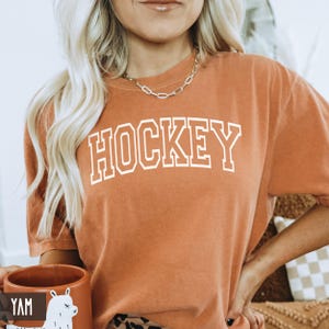 A woman holding a yam orange mug next to an orange yam colored t-shirt with white HOCKEY text.