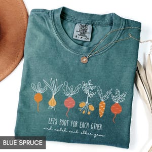 May include: Blue spruce t-shirt with a graphic of root vegetables and the text "LETS ROOT FOR EACH OTHER and watch each other grow." The shirt has a crew neck and short sleeves. A gold necklace with a pendant is on the shirt.