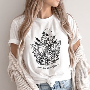 May include: White t-shirt with a black and white illustration of a skeleton holding a book. The text "Just One More Chapter" is printed below the illustration.