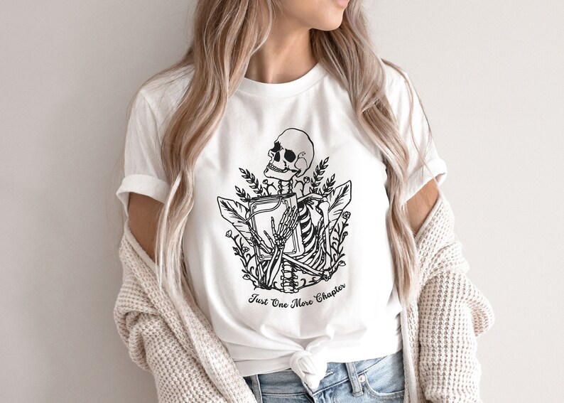 May include: White t-shirt with a black and white illustration of a skeleton holding a book. The text "Just One More Chapter" is printed below the illustration.