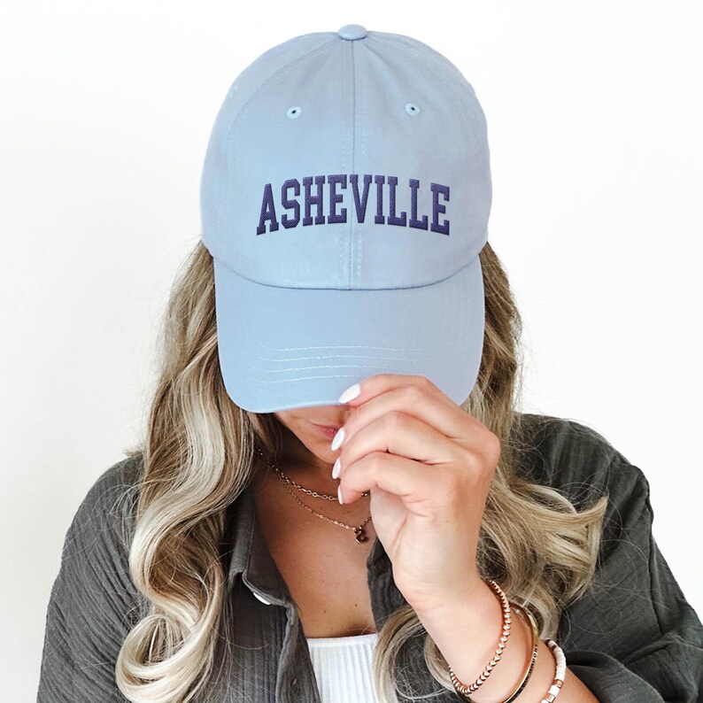 Puede incluir: Gorra de b&eacute;isbol azul claro con la palabra "ASHEVILLE" bordada en azul marino. La gorra est&aacute; hecha de un material suave y tiene una visera curva. Una mujer lleva la gorra.