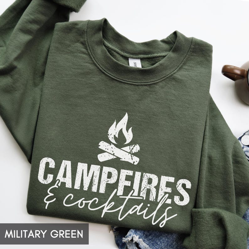 May include: Military green sweatshirt with the words "Campfires & Cocktails" in white script. The design includes a campfire graphic. The sweatshirt is a crew neck style.