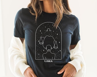 May include: A black short-sleeve t-shirt featuring a white line art graphic of a person with an afro hairstyle holding scales within an arched frame. Above the figure is a Libra zodiac symbol, and below the graphic is the text LIBRA.