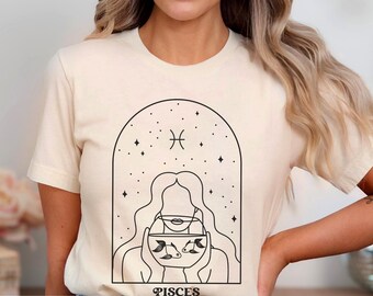May include: A cream-colored short-sleeve t-shirt featuring a black line art graphic of a person holding a fishbowl with two fish inside, framed by an archway with stars and the Pisces zodiac symbol above, with the word PISCES printed below.