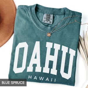 May include: A folded blue spruce t-shirt with the word "OAHU" in large white block letters and "HAWAII" underneath. A gold necklace with a pendant rests on the shirt. The shirt is labeled "Comfort Colors" and is size small.