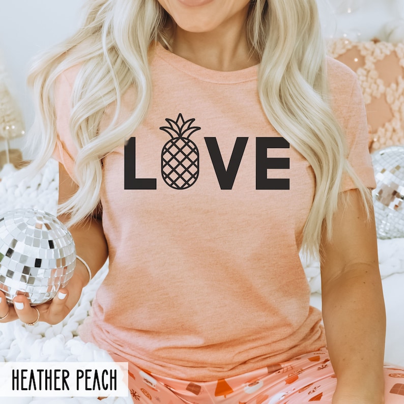 May include: A peach colored t-shirt with the word "LOVE" in black text. A pineapple graphic is in the center of the word "LOVE". The text "HEATHER PEACH" is at the bottom of the image.