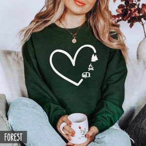 May include: A dark green sweatshirt with a white heart design. The heart includes a campfire, a camper, and a pine tree. The text "FOREST" is visible in the bottom left corner of the image.