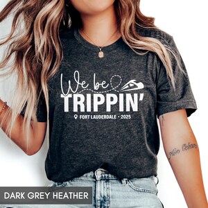 May include: A dark grey heather t-shirt with the text "We be trippin' Fort Lauderdale - 2025" in white.