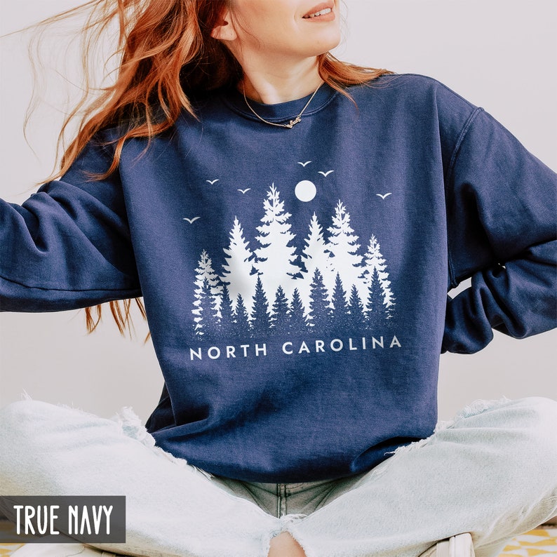 May include: A navy blue sweatshirt with a white graphic of a forest scene with a moon and birds. The text "NORTH CAROLINA" is printed below the graphic.