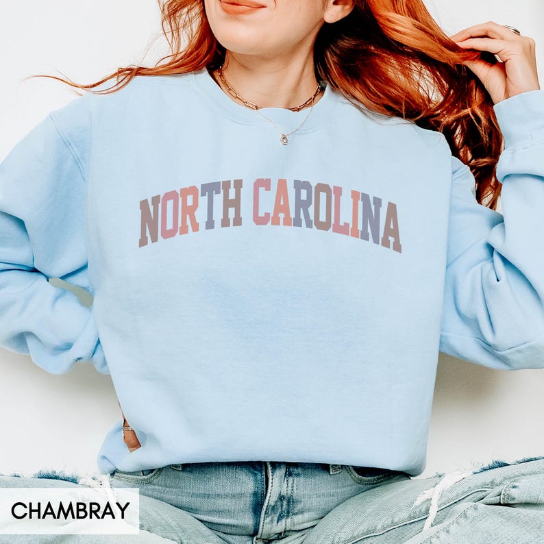 Puede incluir: Una sudadera azul claro con el texto "NORTH CAROLINA" en un dise&ntilde;o multicolor arqueado. La sudadera es de manga larga y cuello redondo.