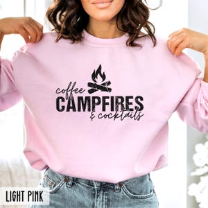 A person wears a light pink crewneck sweatshirt featuring black text that reads coffee CAMPFIRES & cocktails with a campfire graphic.