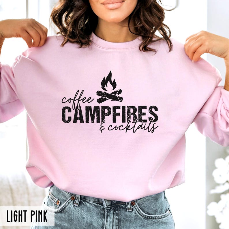 A person wears a light pink crewneck sweatshirt featuring black text that reads coffee CAMPFIRES & cocktails with a campfire graphic.