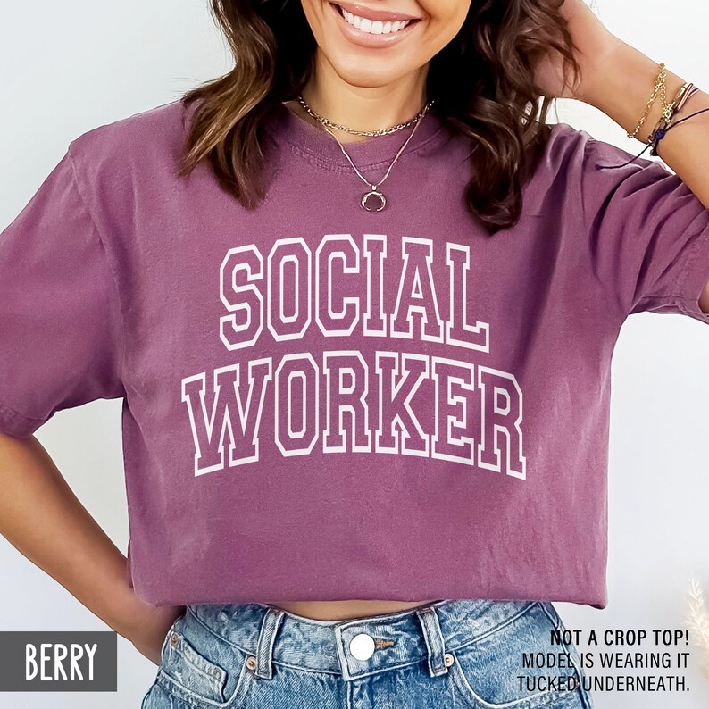 Puede incluir: Una camiseta de color malva con el texto "SOCIAL WORKER" impreso en blanco. El texto est&aacute; en una fuente en negrita y de bloque. La modelo lleva la camiseta metida en unos vaqueros azules.