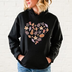 May include: Black hoodie with a heart-shaped design featuring autumn-themed elements like leaves, acorns, mushrooms, and a pumpkin in shades of orange, brown, and pink.