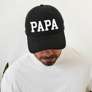 May include: Black baseball cap with the word "PAPA" in white, block letters. The cap is worn by a person wearing a white shirt. The cap has a curved brim and a button on top.
