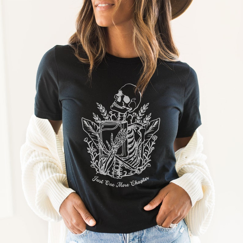 May include: Black t-shirt with a white graphic of a skeleton holding a book and surrounded by flowers. The text "Just One More Chapter" is printed below the graphic.