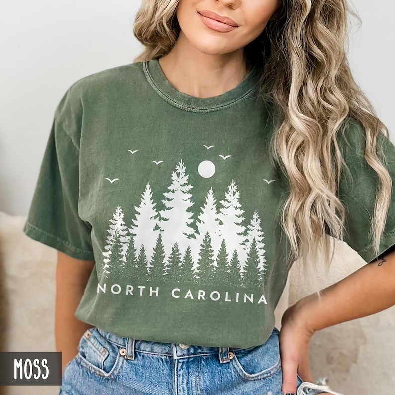 以下が含まれることがあります： 白い森のシルエットと「NORTH CAROLINA」の文字が書かれた緑色のTシャツです。
