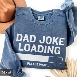 May include: Denim blue sweatshirt with the text "DAD JOKE LOADING" in white, along with a loading bar graphic and the words "PLEASE WAIT...". The sweatshirt is a Comfort Colors brand.