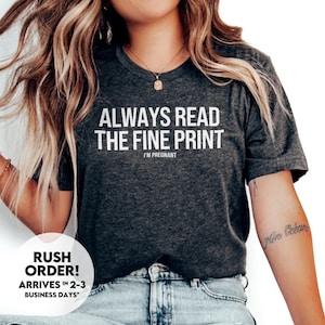 May include: A dark gray t-shirt with the text "ALWAYS READ THE FINE PRINT I'M PREGNANT" in white. The shirt is being worn by a person with long blonde hair and blue jeans.