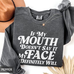 Puede incluir: Una sudadera gris oscuro con el texto "If My Mouth Doesn't Say It My Face Definitely Will" en blanco. Tambi&eacute;n se ven un collar dorado y un sombrero marr&oacute;n.