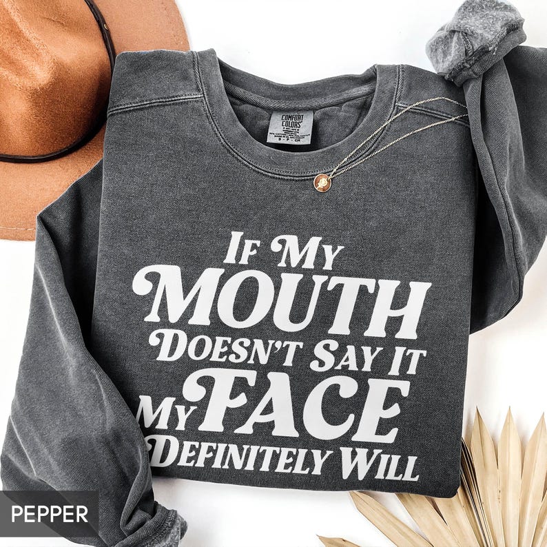 Puede incluir: Una sudadera gris oscuro con el texto "If My Mouth Doesn't Say It My Face Definitely Will" en blanco. Tambi&eacute;n se ven un collar dorado y un sombrero marr&oacute;n.