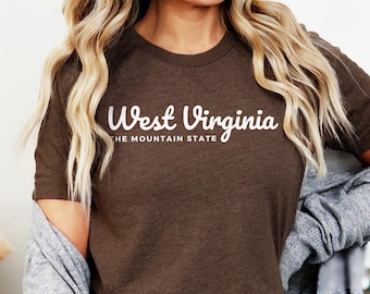 Puede incluir: Una camiseta de manga corta en color marrón jaspeado con el texto blanco West Virginia THE MOUNTAIN STATE impreso en el pecho. La camiseta tiene un cuello redondo y una apariencia de tela suave y texturizada con acabado jaspeado.