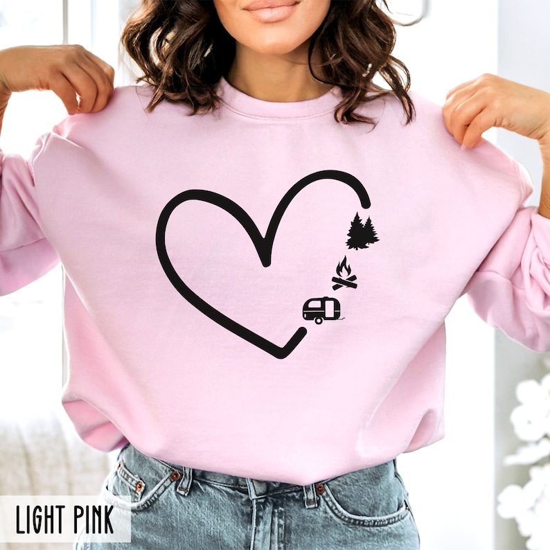 May include: A light pink sweatshirt with a black heart design. The heart is filled with a camper, a campfire, and trees. The text "LIGHT PINK" is at the bottom of the image.