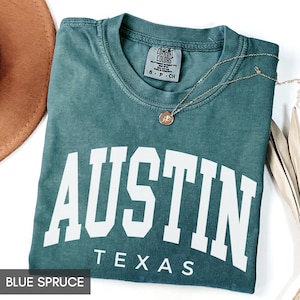May include: Blue spruce t-shirt with the word "AUSTIN" in large white letters and "TEXAS" underneath. The shirt has a crew neck and short sleeves. A gold necklace with a pendant is draped across the shirt.