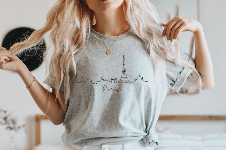 Op de afbeelding: Een grijs T-shirt met een zwarte lijntekening van de Eiffeltoren en het woord "Paris" eronder.