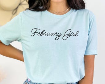 May include: A light blue short-sleeve t-shirt featuring the text "February Girl" printed in a black cursive script across the chest. The garment is made of a heathered fabric and is paired with blue denim jeans, showcasing a casual style.