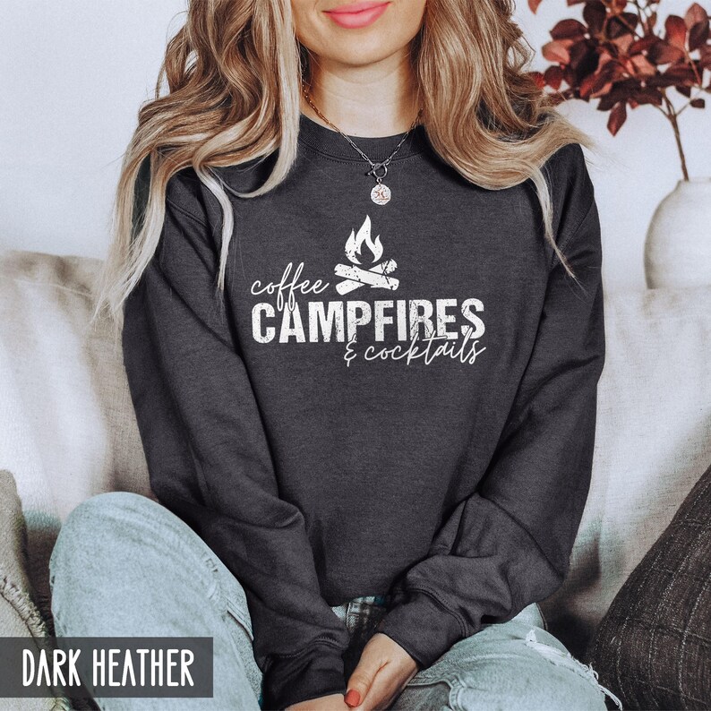 A person wears a dark heather gray crewneck sweatshirt featuring white text that reads coffee CAMPFIRES & cocktails with a campfire graphic.