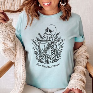 May include: A light blue t-shirt with a black and white graphic of a skeleton holding a book. The text "Just One More Chapter" is printed below the graphic.