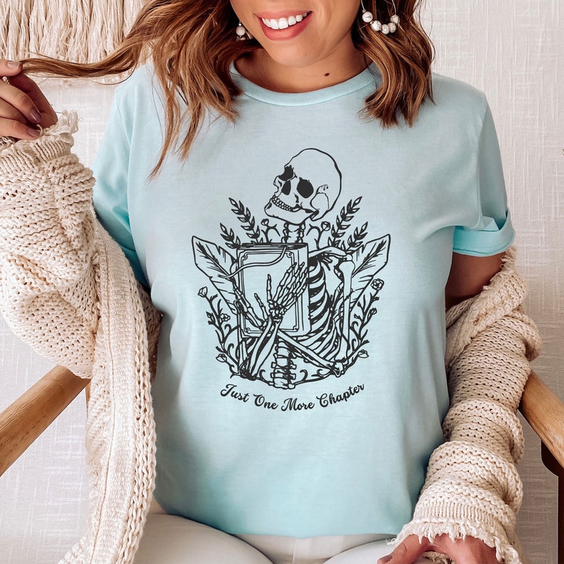 May include: A light blue t-shirt with a black and white graphic of a skeleton holding a book. The text "Just One More Chapter" is printed below the graphic.