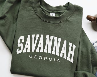 May include: A military green sweatshirt with the word "SAVANNAH" in large white letters arched across the chest, with "GEORGIA" below. The sweatshirt is folded, with a portion of denim jeans visible.