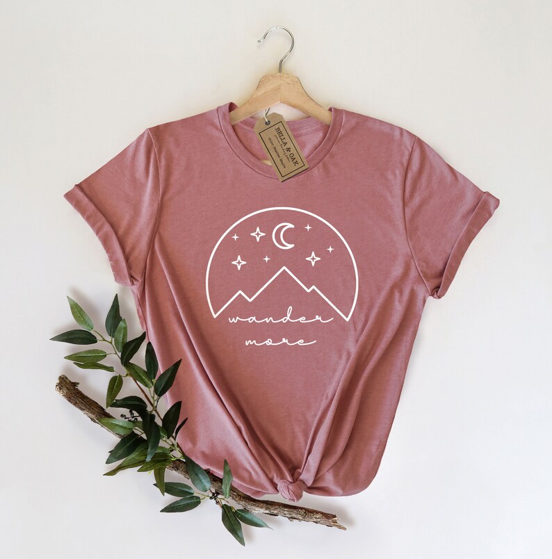 Pode incluir: Uma camiseta rosa p&oacute; com um gr&aacute;fico branco de montanhas, uma lua crescente e estrelas. O texto "wander more" est&aacute; abaixo do gr&aacute;fico.